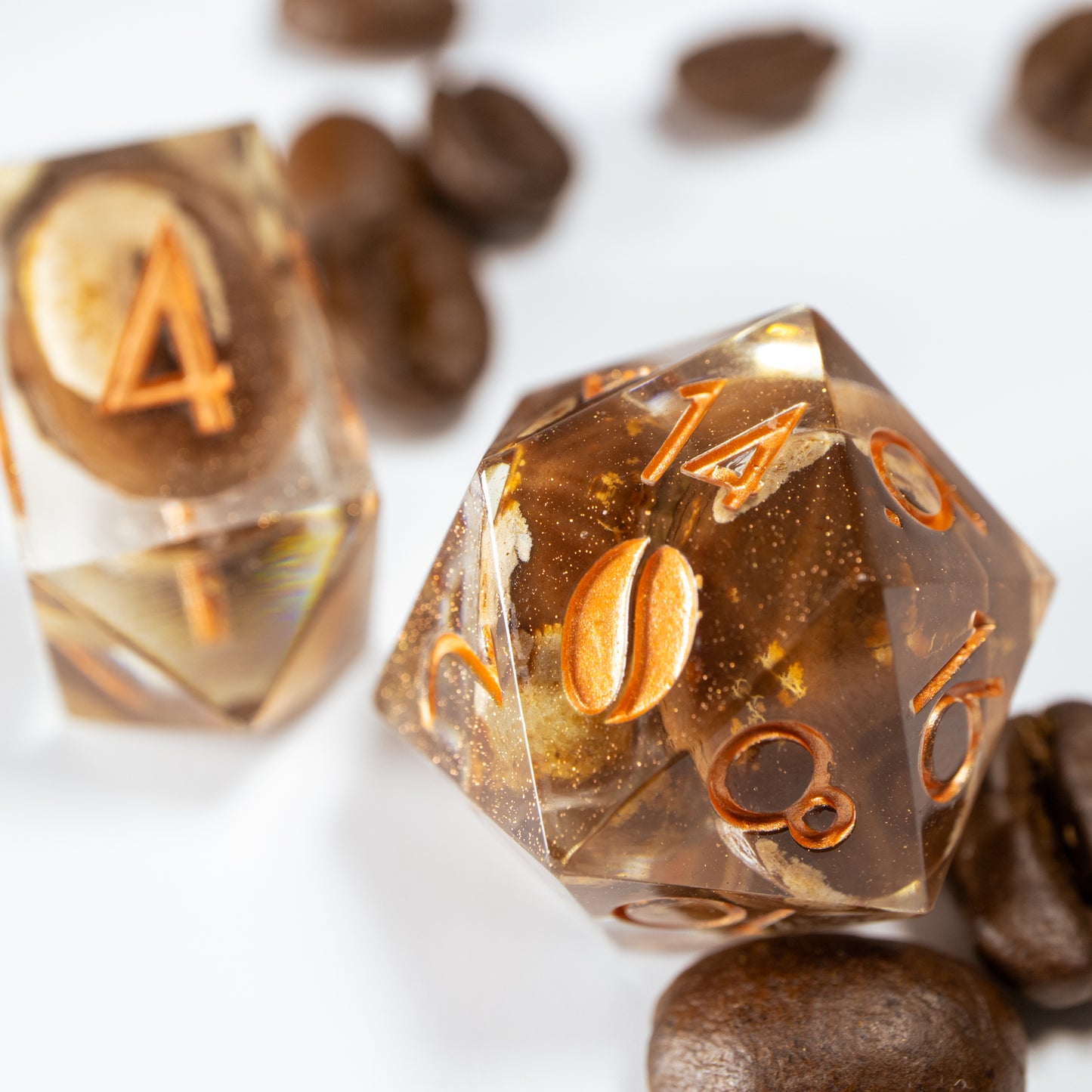 A close-up of a coffee themed twenty sided die. Behind it lies an elongated, crystal-shaped four-sided die, among a scattering of decorative coffee beans.