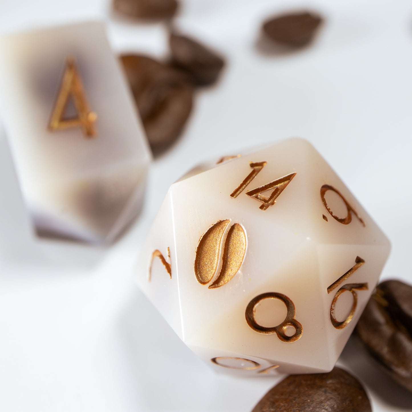 A close-up of a coffee themed twenty sided die. Behind it lies an elongated, crystal-shaped four-sided die, among a scattering of decorative coffee beans.