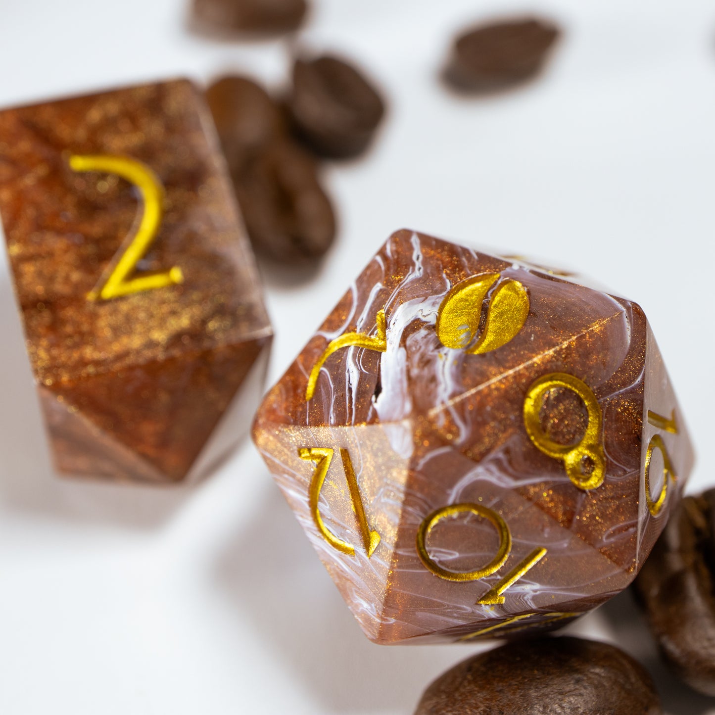 A close-up of a coffee themed twenty sided die. Behind it lies an elongated, crystal-shaped four-sided die, among a scattering of decorative coffee beans.
