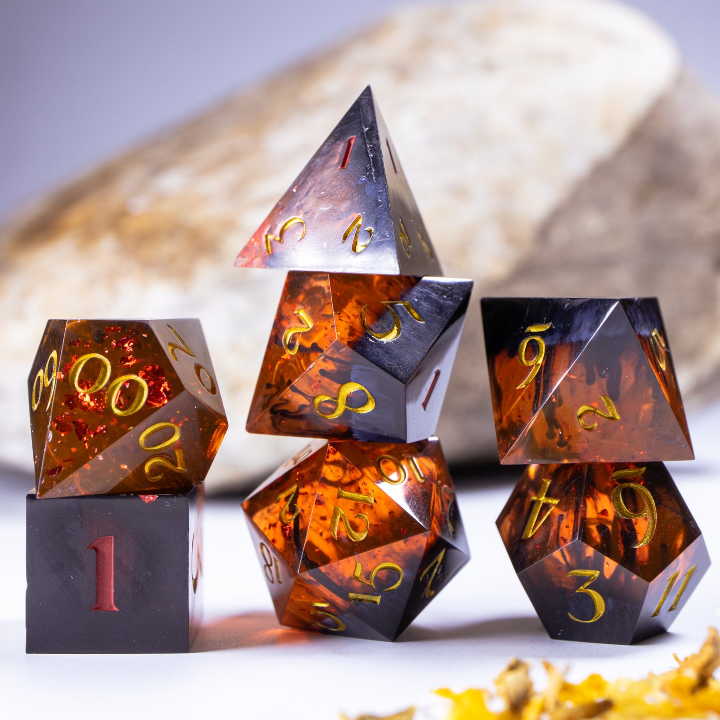 Three towers of stacked polyhedral dice with added background elements, such as an out of focus rock for added texture and decorative leaves.