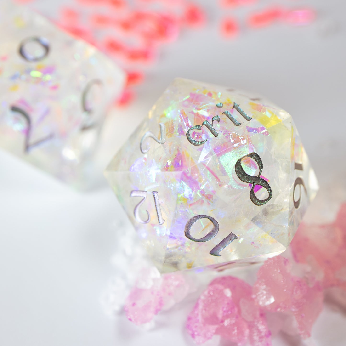 A close-up of a twenty sided die with iridescent mylar flakes. Behind it lies a ten sided die, among a scattering of tiny decorative pink shards.