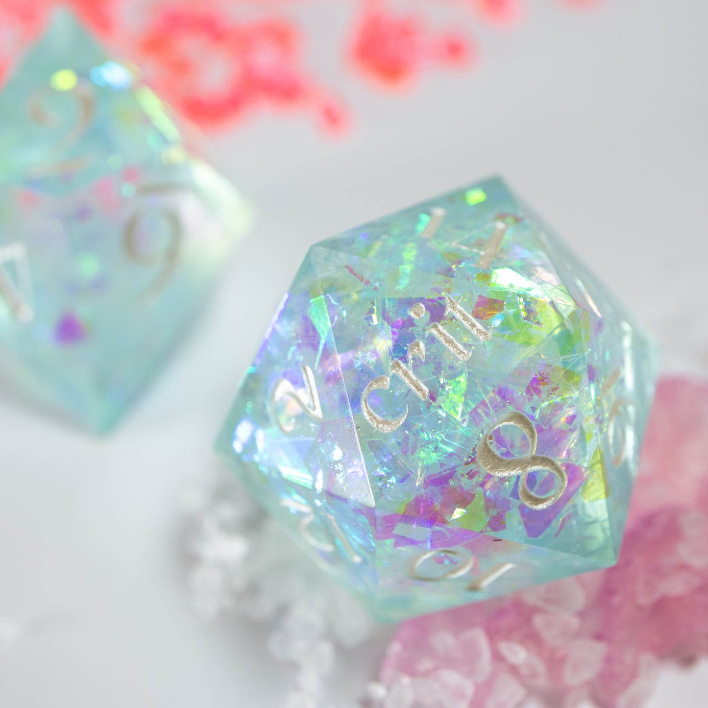 A close-up of a light blue twenty sided die iridescent mylar flakes. Behind it lies a ten sided die, among a scattering of tiny decorative pink shards.