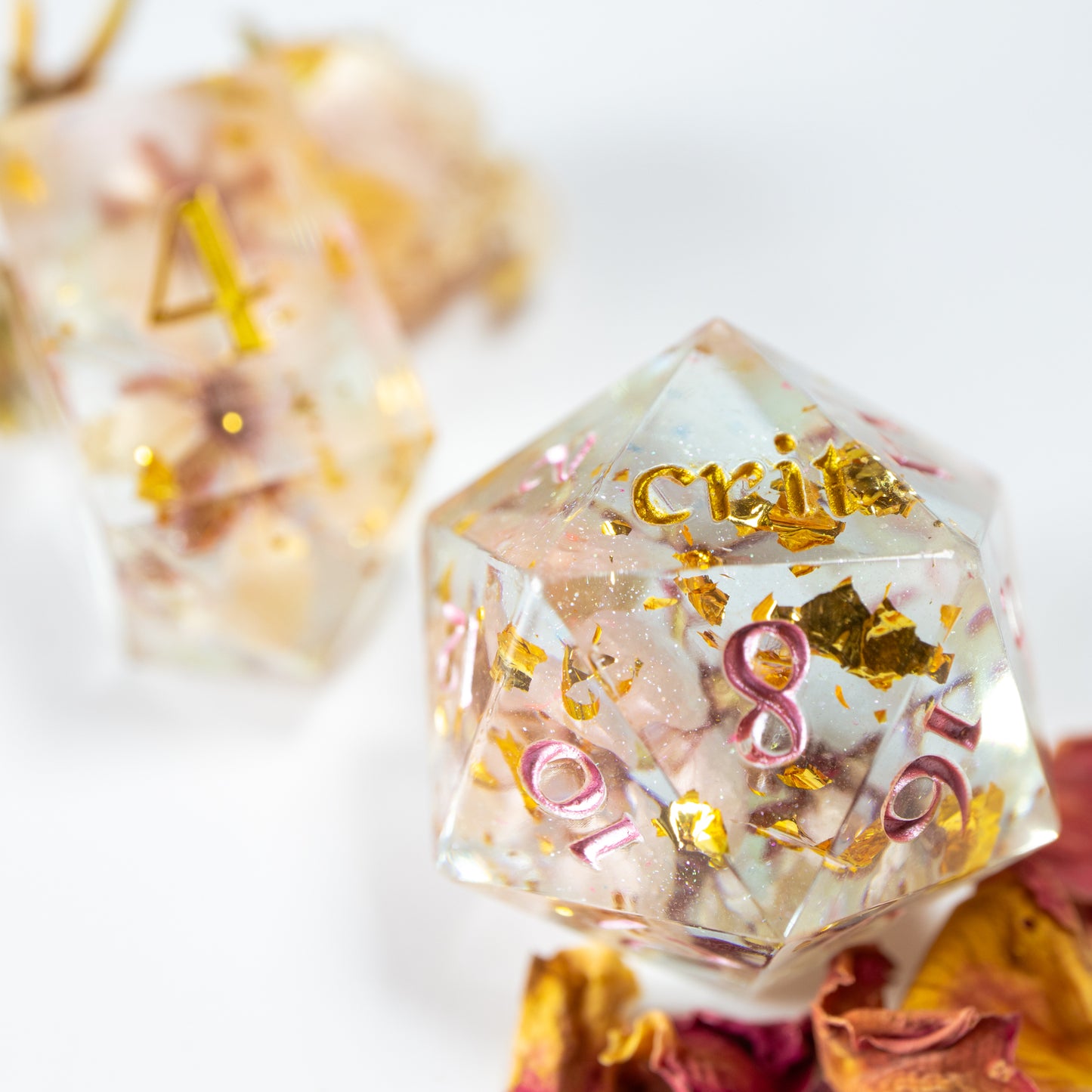 A close-up of a twenty sided die with gold foil flecks and dried cherry blossoms within. Behind it lies a ten sided die, among a scattering of decorative petals.