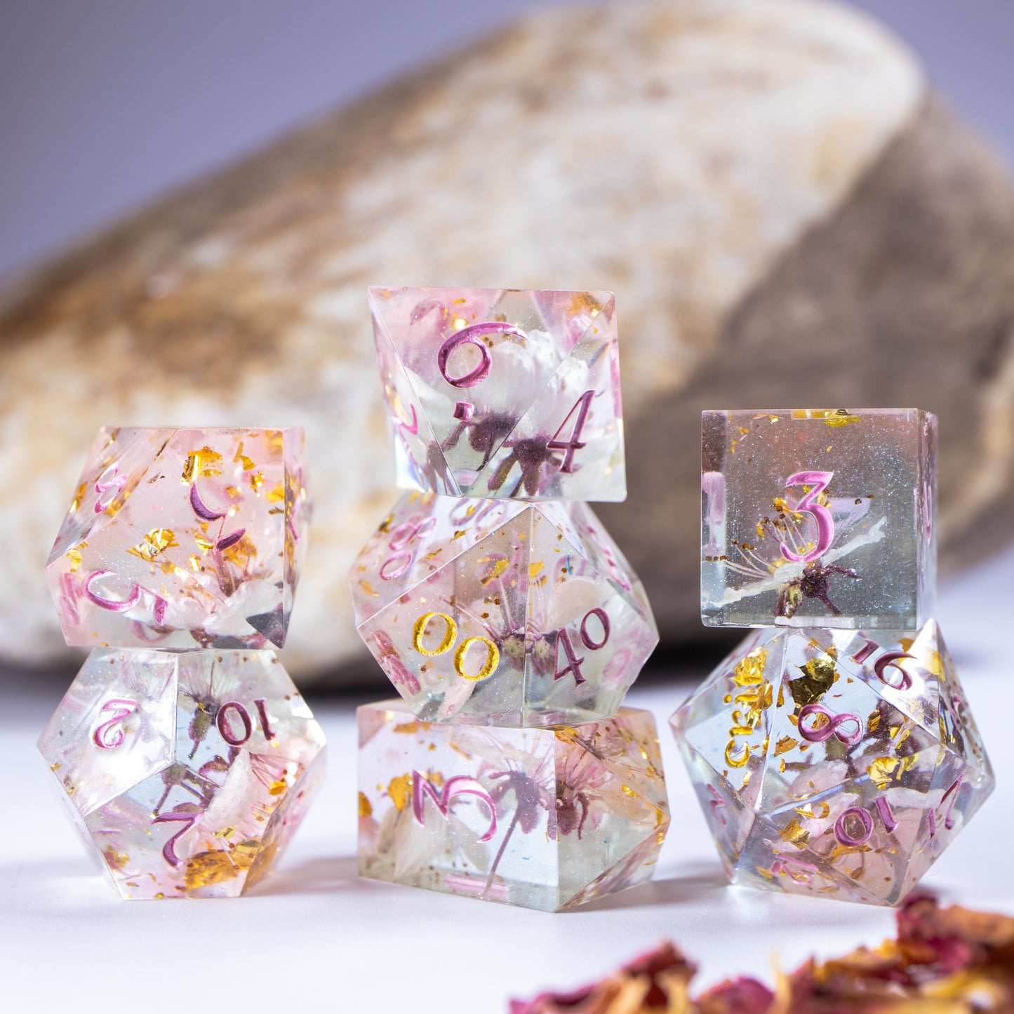 Three towers of stacked polyhedral dice with added background elements, such as an out of focus rock for added texture and decorative petals.