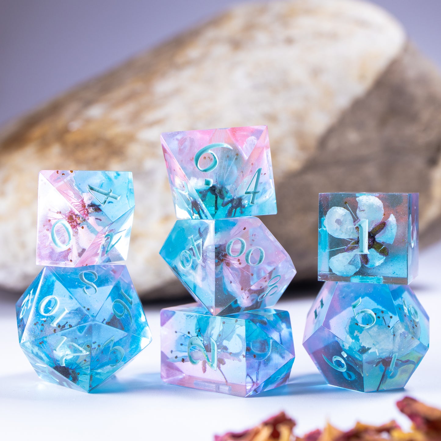 Three towers of stacked polyhedral dice with added background elements, such as an out of focus rock for added texture and decorative petals.