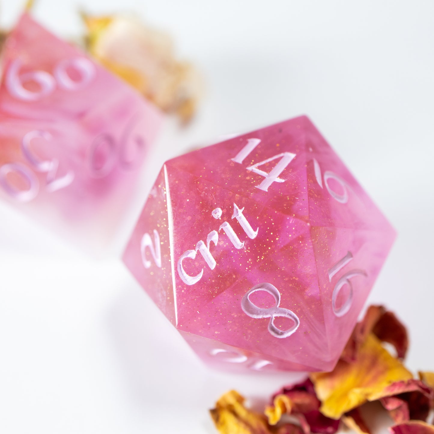 A close-up of a light pink twenty sided die iridescent mylar flakes. Behind it lies a ten sided die, among a scattering of decorative leaves.