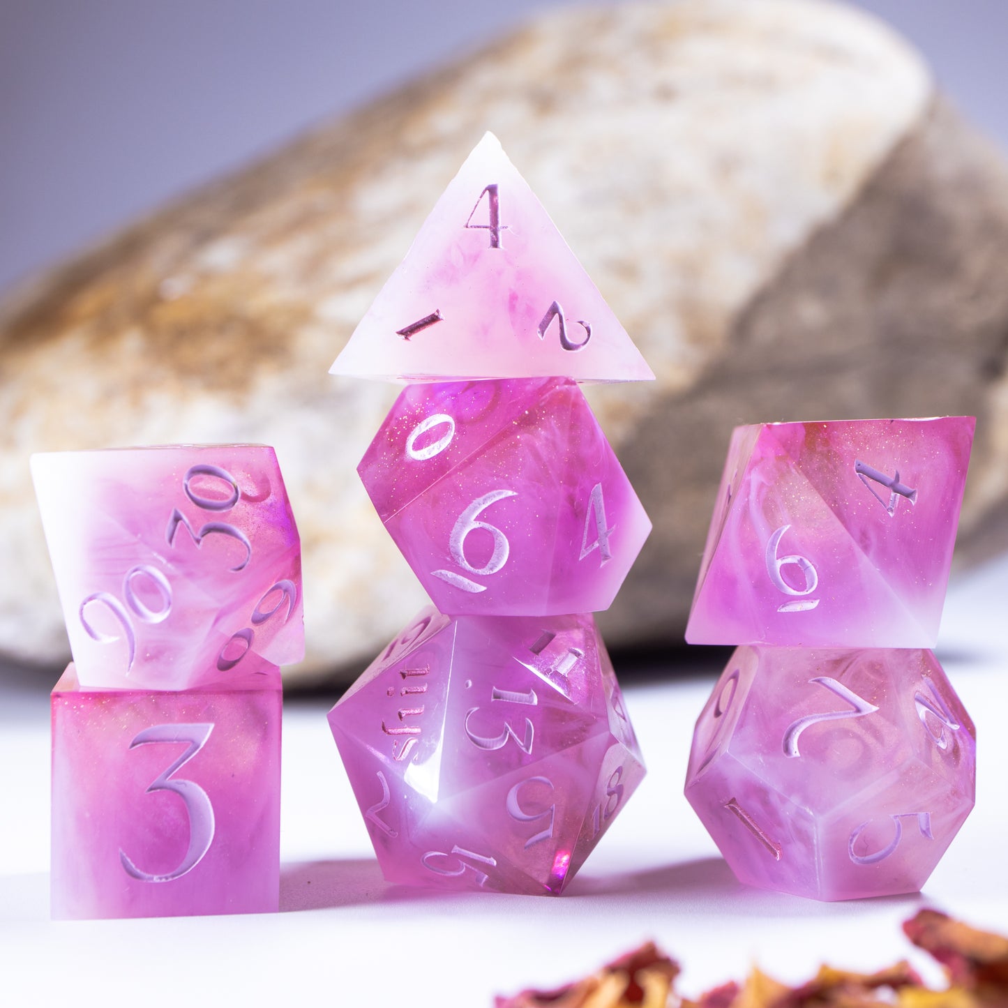 Three towers of stacked polyhedral dice with added background elements, such as an out of focus rock for added texture and decorative leaves and petals.