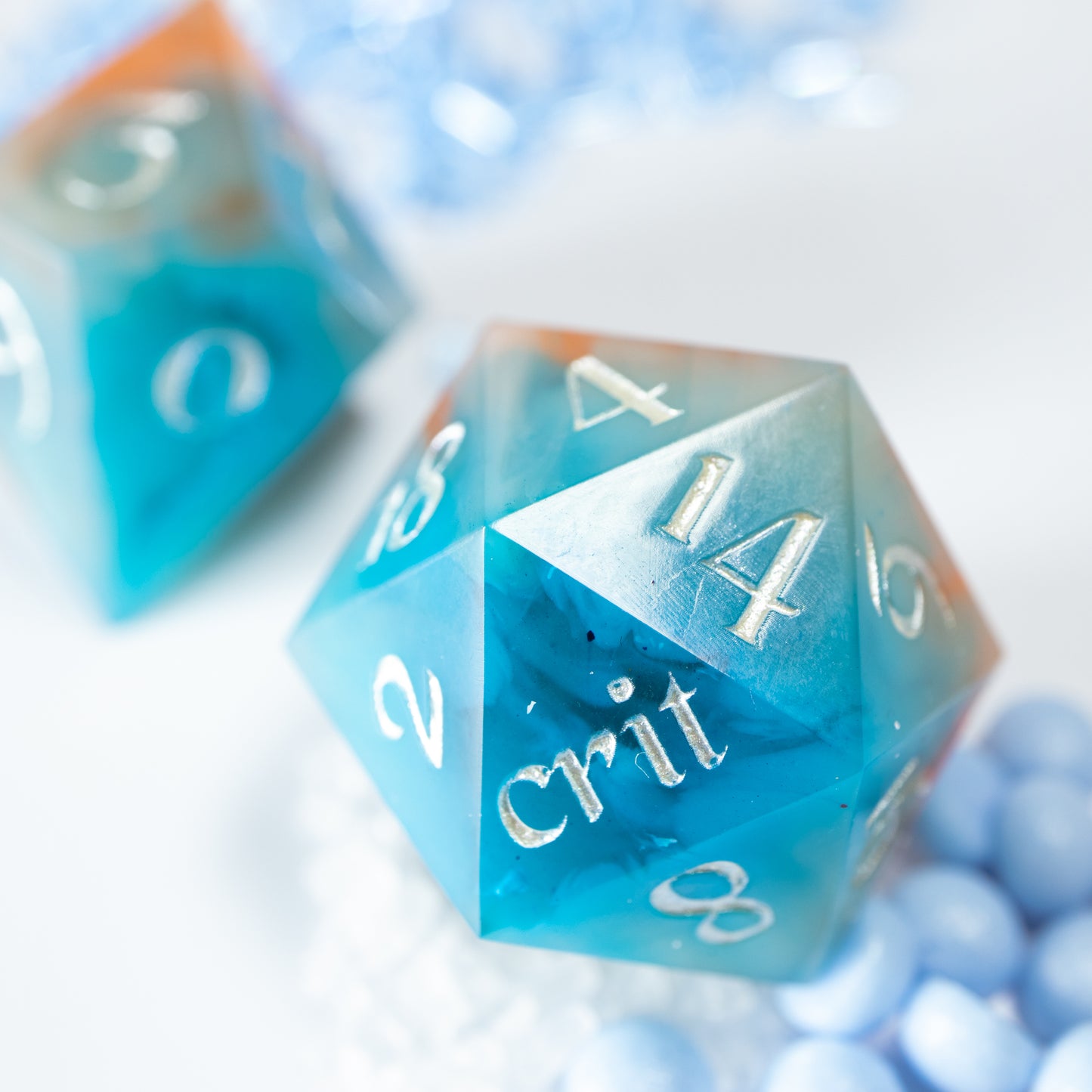 A close-up of a teal coloured twenty sided die. Behind it lies a ten sided die, among a scattering of tiny decorative blue wax pearls.
