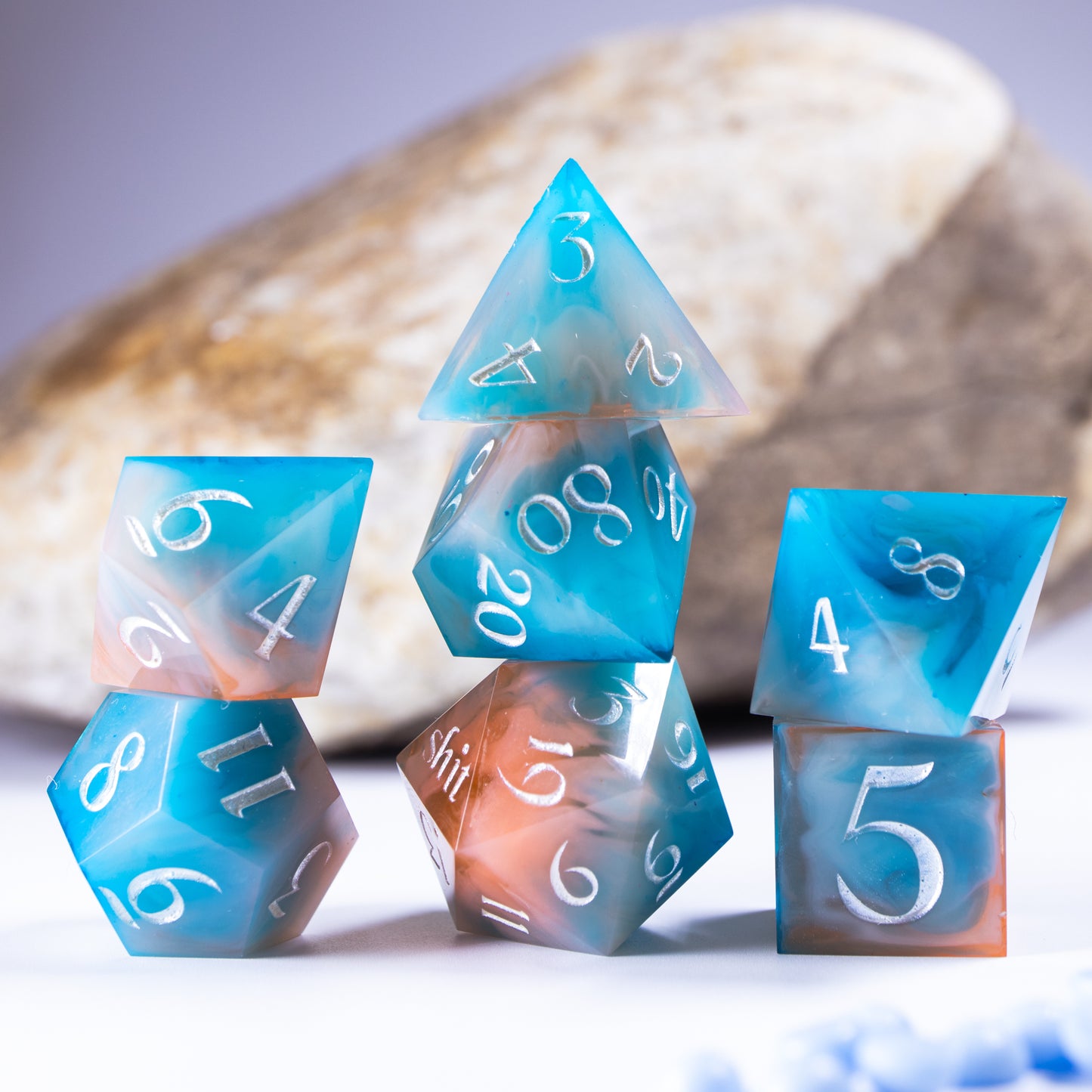 Three towers of stacked polyhedral dice with added background elements, such as an out of focus rock for added texture and tiny decorative wax pearls.