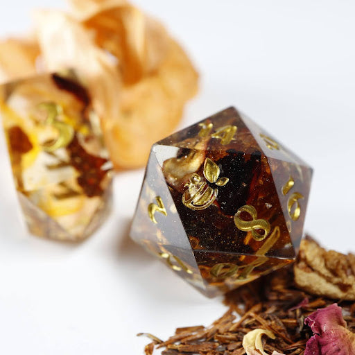 A close-up of a tea themed twenty sided die. Behind it lies an elongated, crystal-shaped four-sided die, among a scattering of decorative tea leaves.