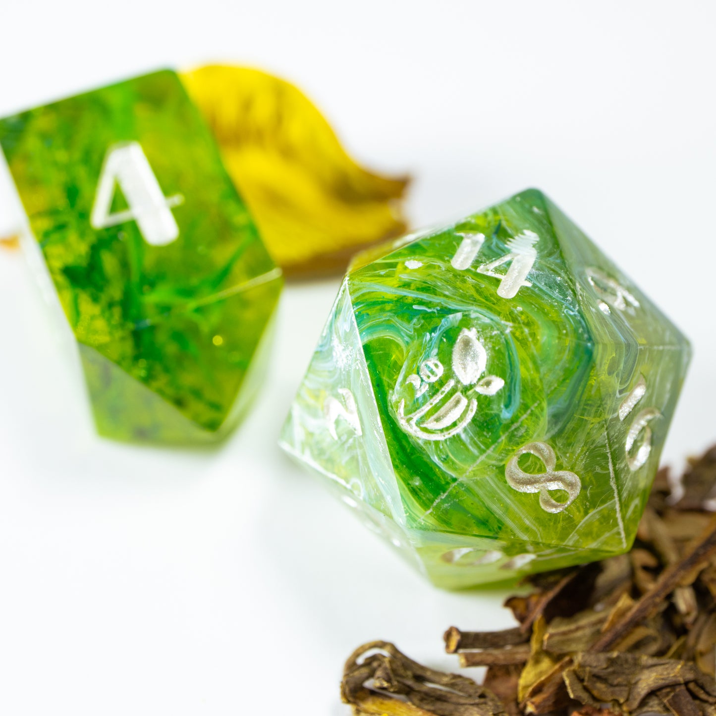 A close-up of a tea themed twenty sided die. Behind it lies an elongated, crystal-shaped four-sided die, among a scattering of decorative tea leaves.