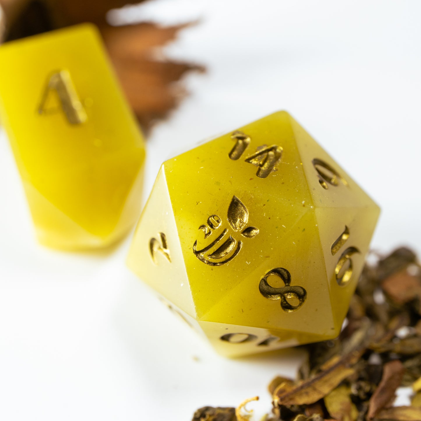A close-up of a tea themed twenty sided die. Behind it lies an elongated, crystal-shaped four-sided die, among a scattering of decorative tea leaves.