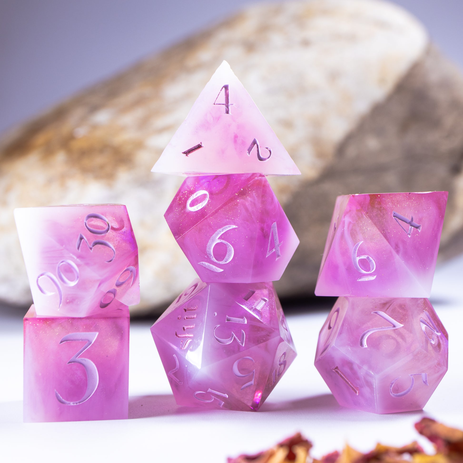 Three towers of stacked polyhedral dice with added background elements, such as an out of focus rock for added texture and decorative leaves and petals.